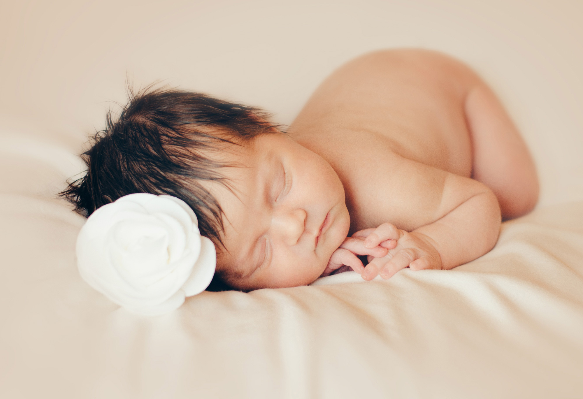 Sleeping asian baby girl on a smooth beige sheet. Baby has a white rose on her head.