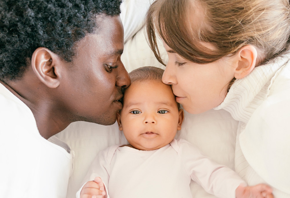 Mixed race family photo. Baby is at the center, mom and dad on each side kissing the baby's forehead.